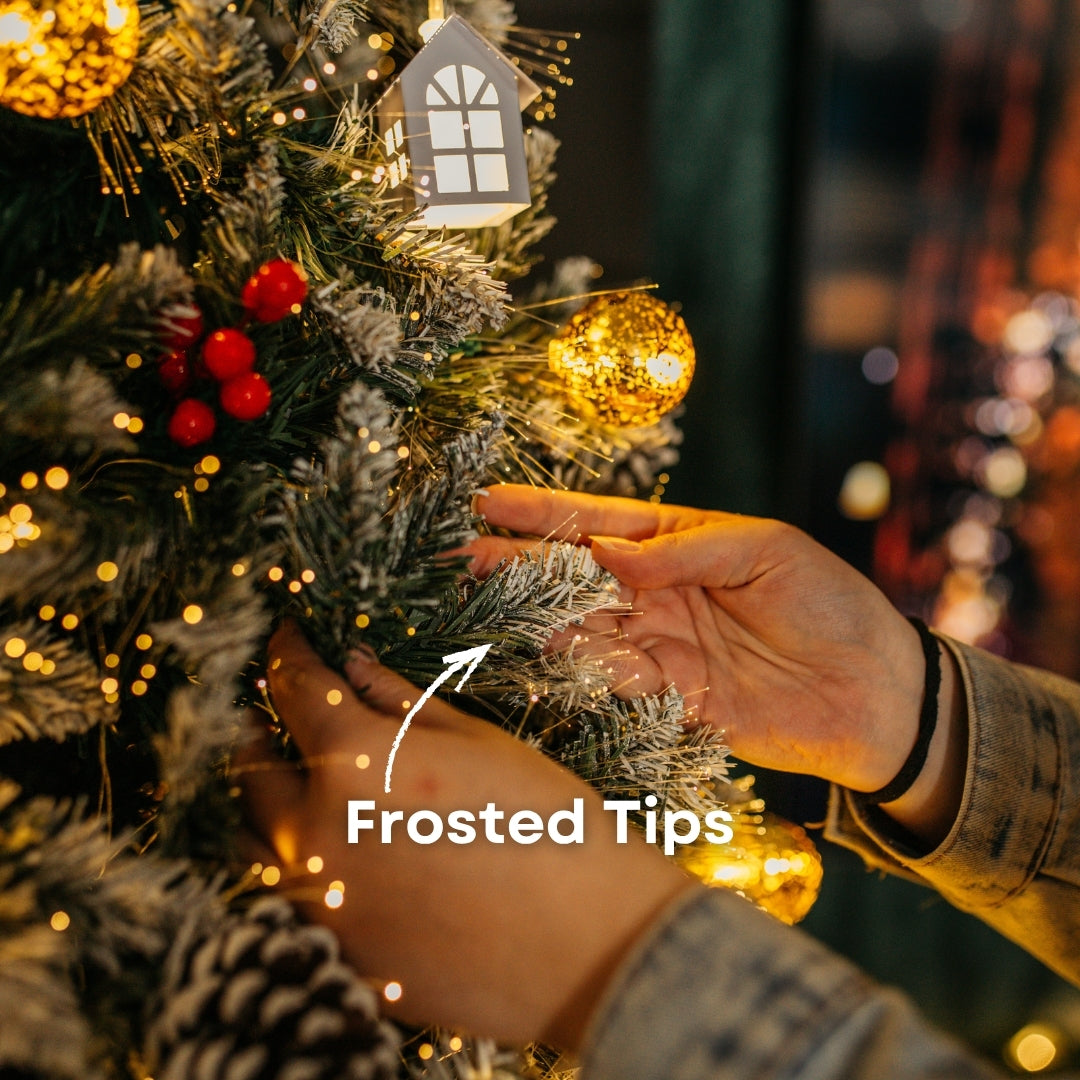 Hands touching the frosted branch tips of a Christmas tree beside an illuminated mini house and golden bauble