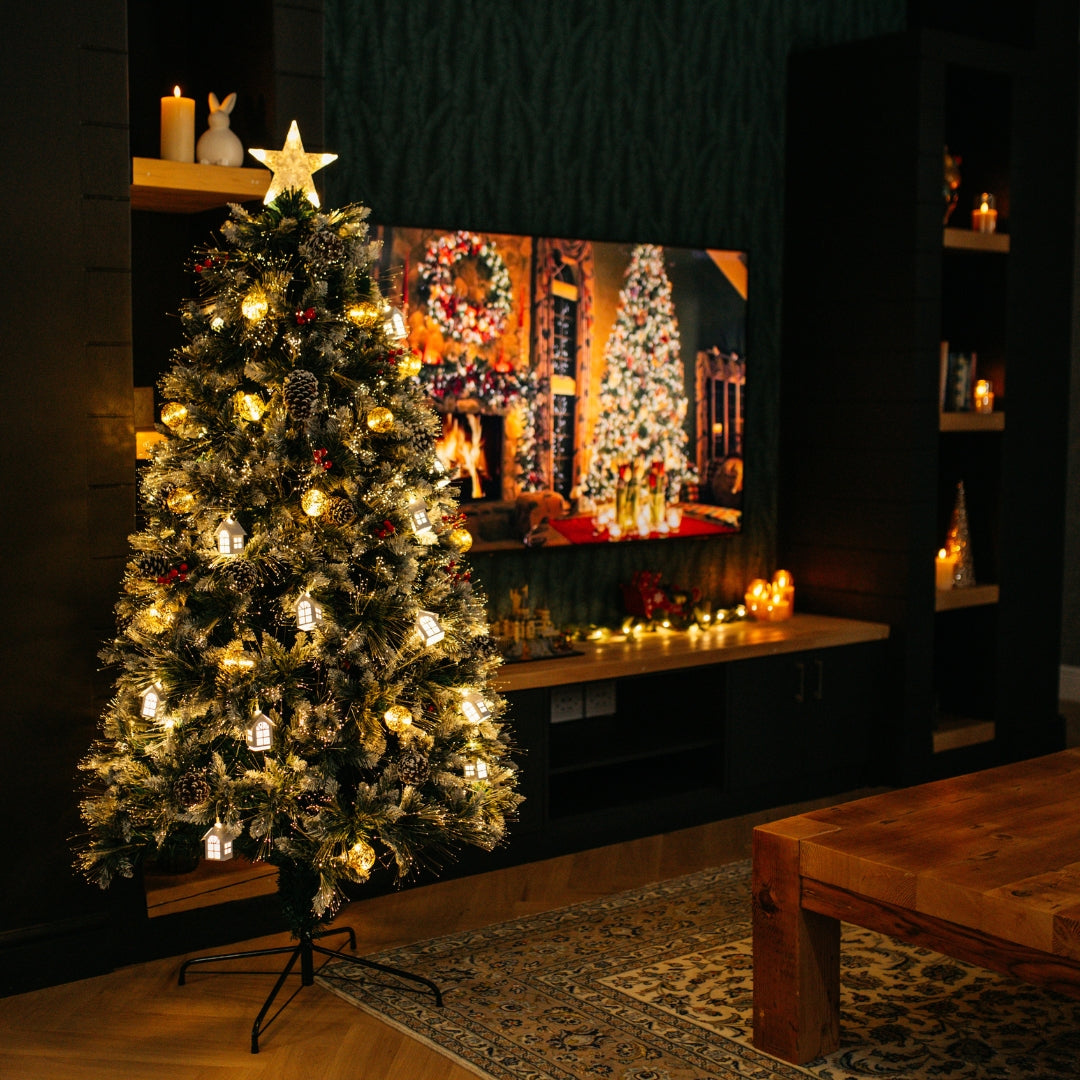 1.8m prelit Christmas tree with warm white LEDs glowing in a dark living room beside a TV displaying a fireplace Christmas scene