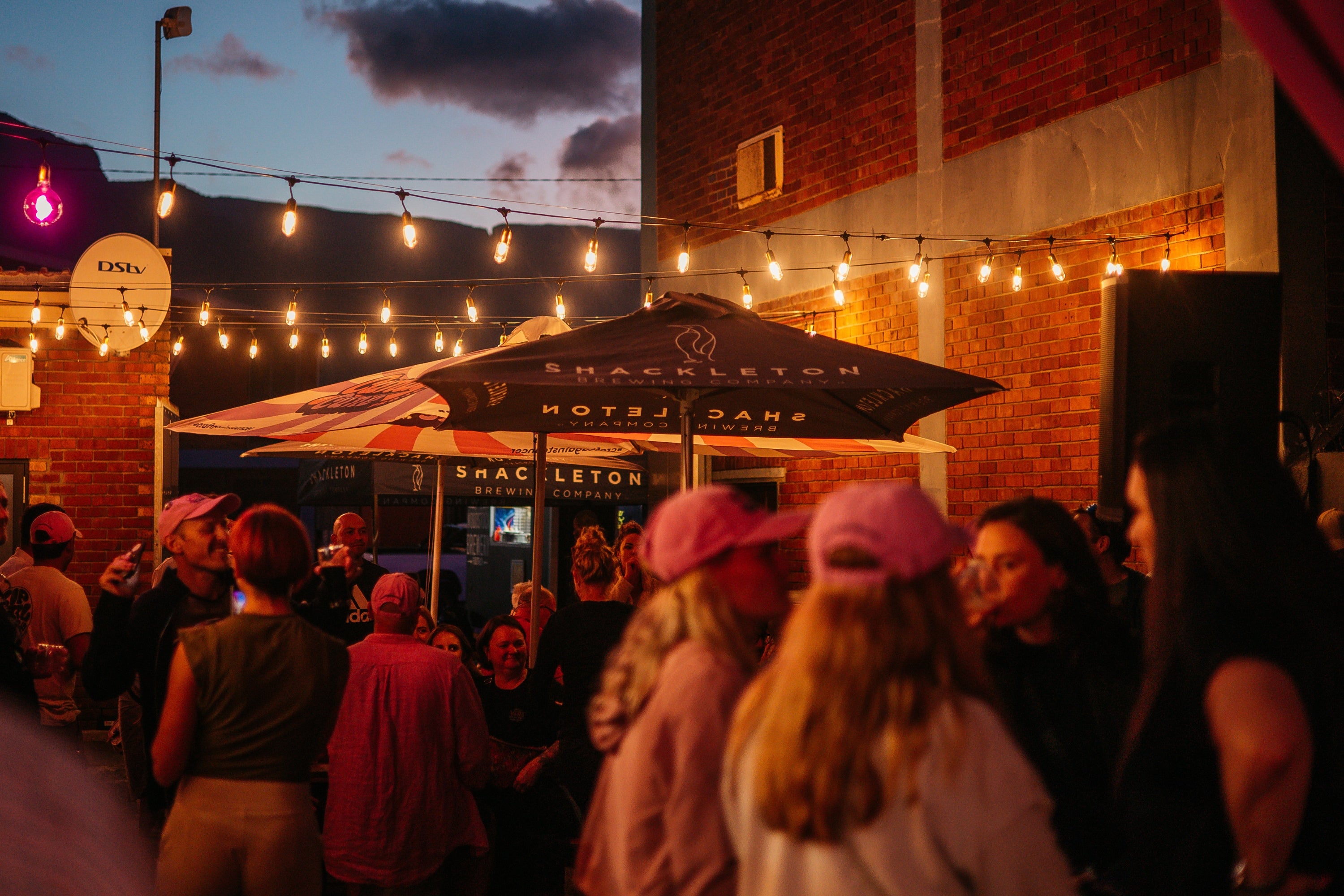 A crowd of people gathered outdoors at dusk under festoon string lights in front of a brick building. A large patio umbrella with the words 'SHACKLETON' are visible.
