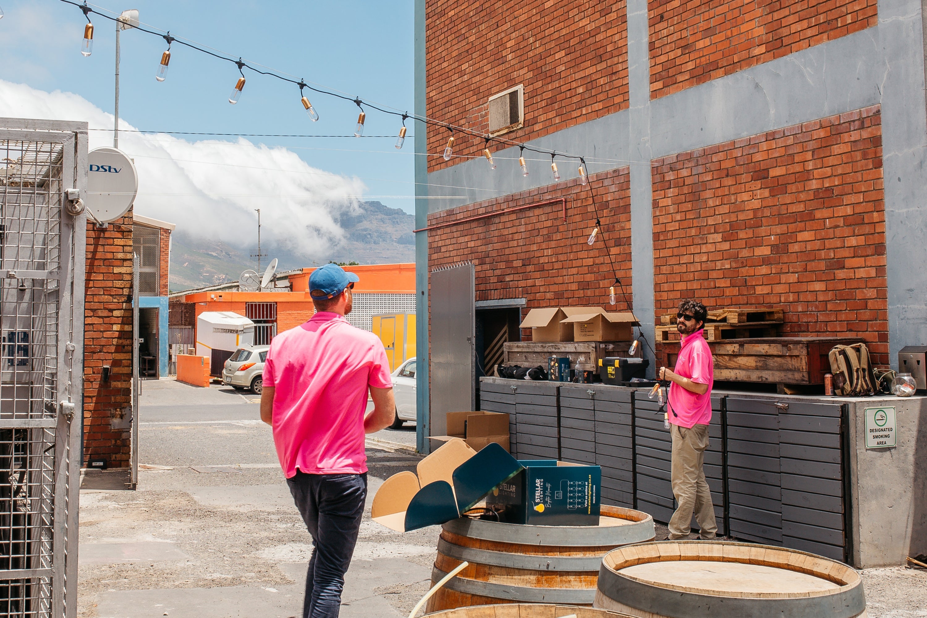 Two people wearing matching bright pink Stellar Lighting shirts work outdoors in a brickyard or warehouse setting, setting up festoon lights. The scene includes a red brick building, string lights, mountains in the distance, and a partly cloudy sky.