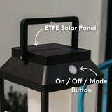 Black outdoor solar glow lantern light fixture with ETFE solar panel and on/off mode button, blurred background