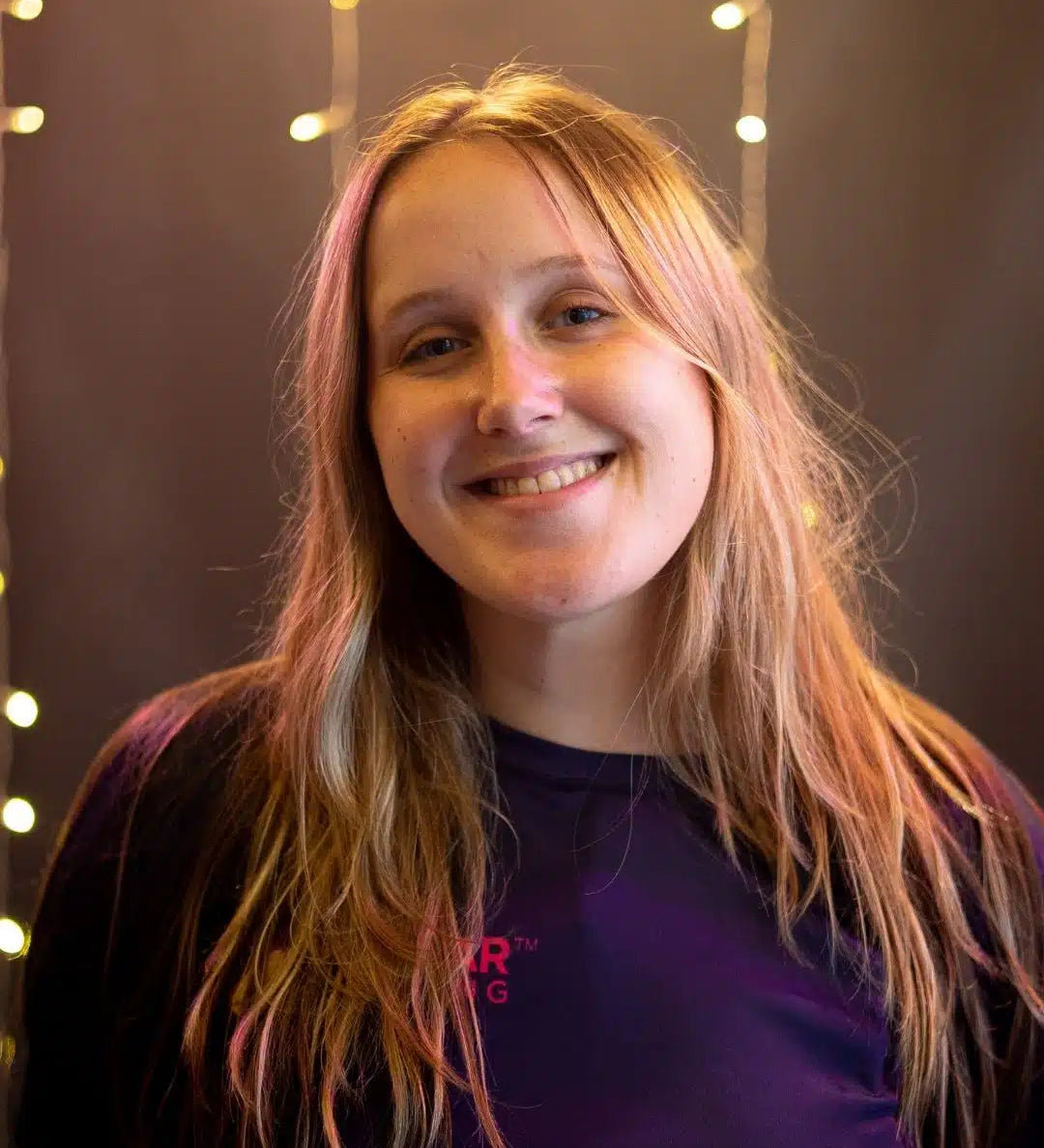 Person with long blonde hair wearing a blue shirt with Stellar Lighting logo, against a dark background with string lights.