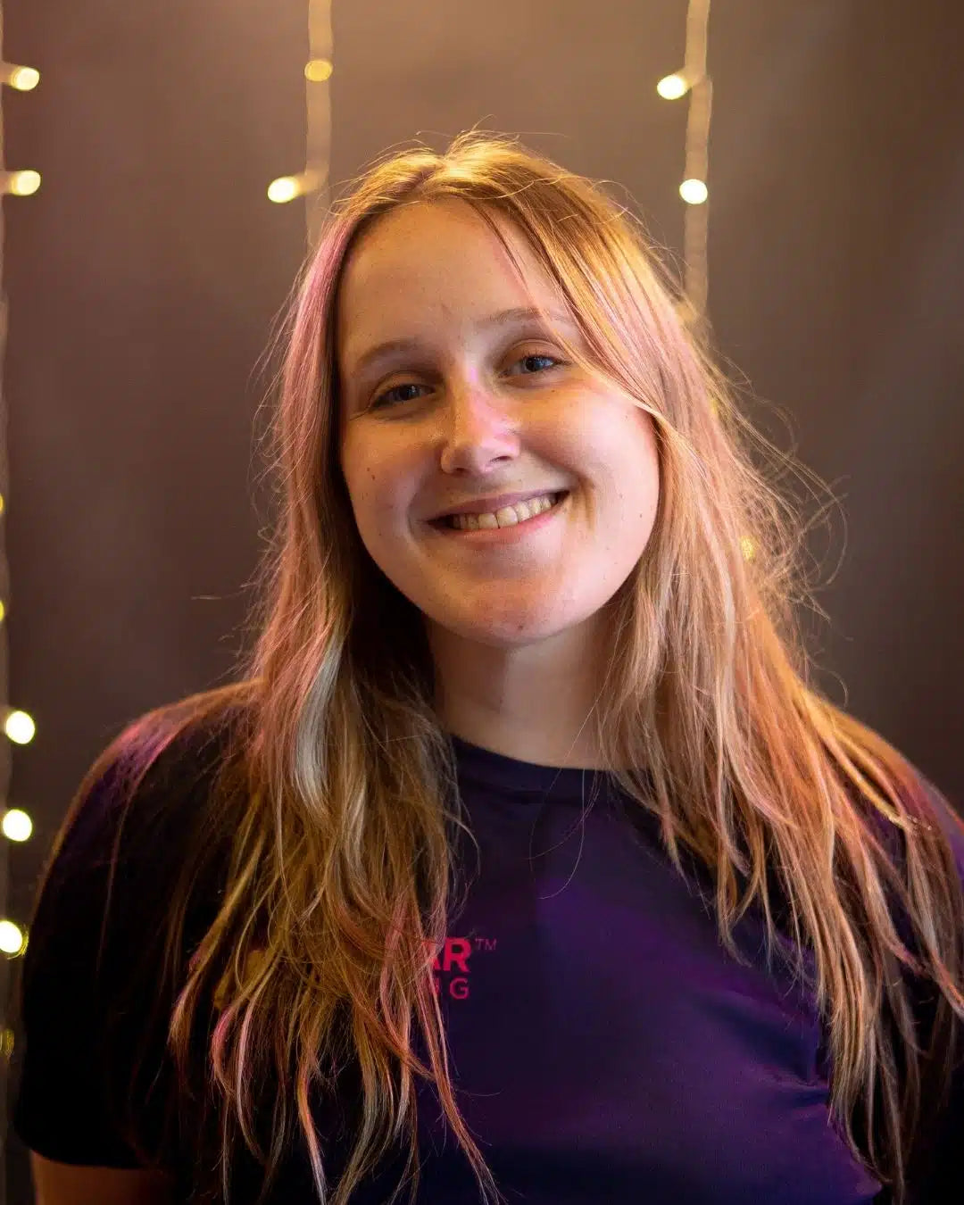 Person with long blonde hair wearing a blue shirt with Stellar Lighting logo, against a dark background with string lights.