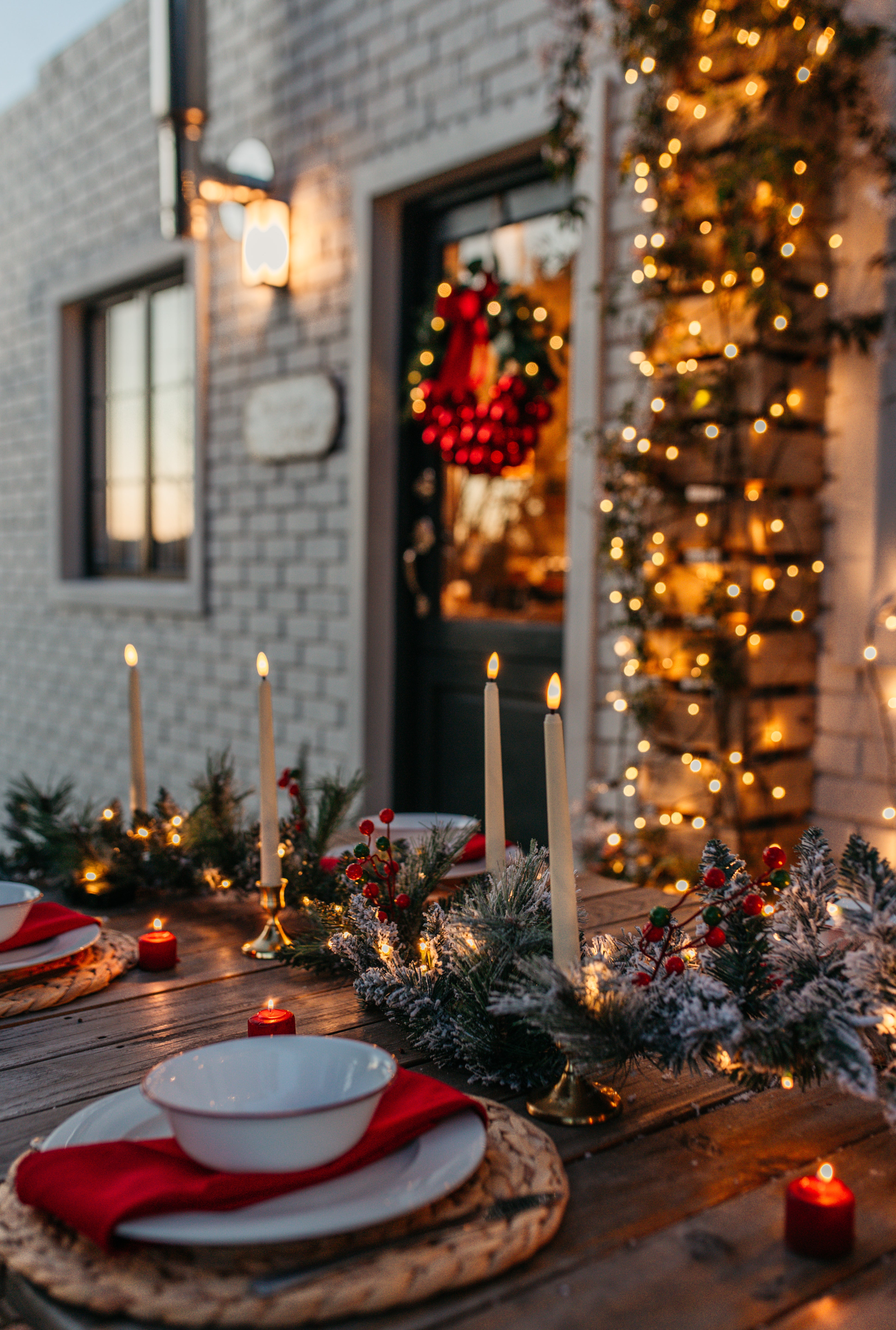 A festive outdoor holiday table setting in front of a brick house, decorated with a green and white frosted garland, white taper candles, red napkins, white plates, and a red berry wreath on the door.