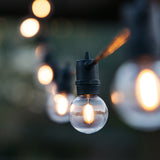 Close-up of a warm white G40 globe bulb on black cable hanging outdoors against a soft green bokeh background