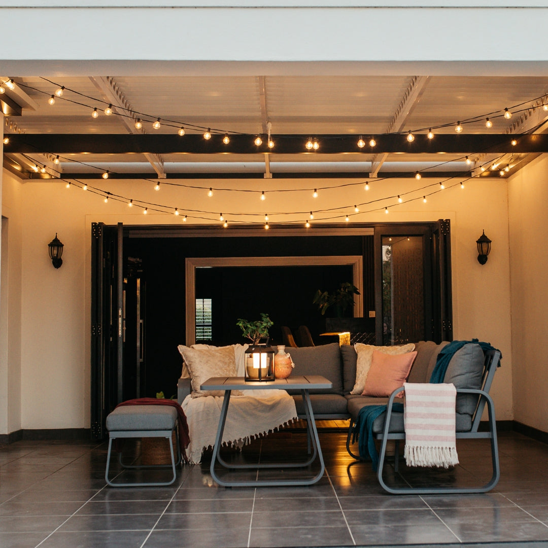Classic String Lights Pro strung across a covered outdoor patio ceiling at night with lounge furniture, a lit braai, and candles creating a warm inviting atmosphere