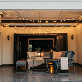 Classic String Lights Pro strung across a covered outdoor patio ceiling at night with lounge furniture, a lit braai, and candles creating a warm inviting atmosphere