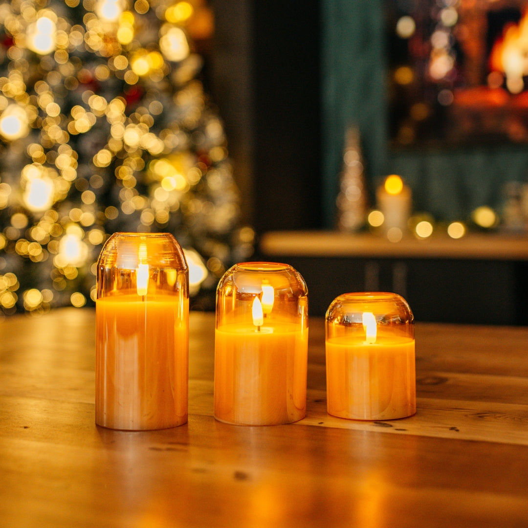 Set of three domed gold glass flameless LED candles glowing on a wooden table with a Christmas tree and fireplace in the background