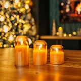 Set of three domed gold glass flameless LED candles glowing on a wooden table with a Christmas tree and fireplace in the background