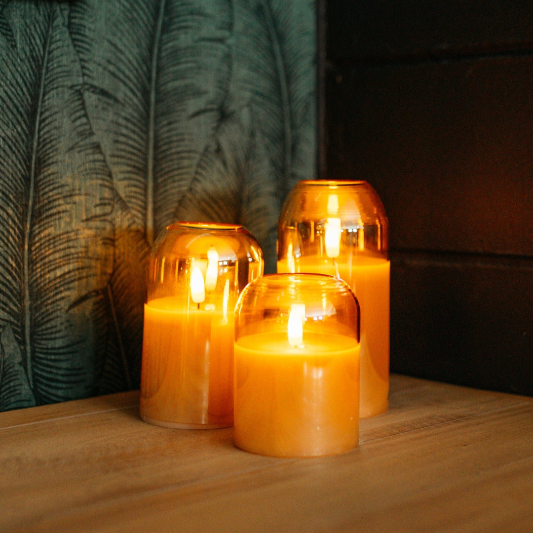 Set of three domed gold glass flameless LED candles glowing on a wooden surface against a dark tropical leaf wallpaper background