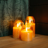 Set of three domed gold glass flameless LED candles glowing on a wooden surface against a dark tropical leaf wallpaper background