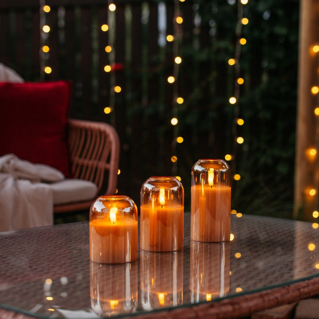 Set of three domed gold glass flameless LED candles glowing on an outdoor glass table with fairy lights in the background