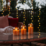 Set of three domed gold glass flameless LED candles glowing on an outdoor glass table at dusk with curtain fairy lights in the background