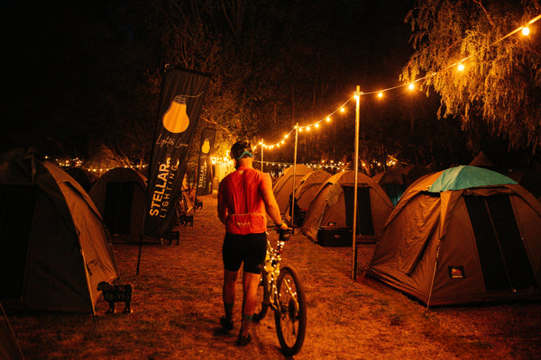 A person standing with a bicycle at night in a camping site, surrounded by tents and warm string lights. A vertical black banner on the left reads 'STELLAR LIGHTS'.
