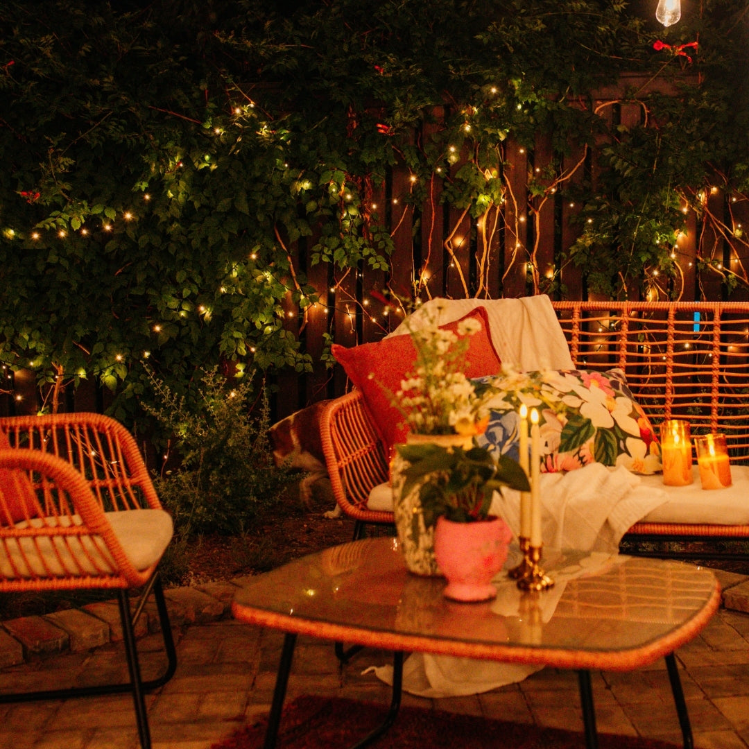 Outdoor evening setting with fairy lights, a table with plants and candles, and a bench with cushions.