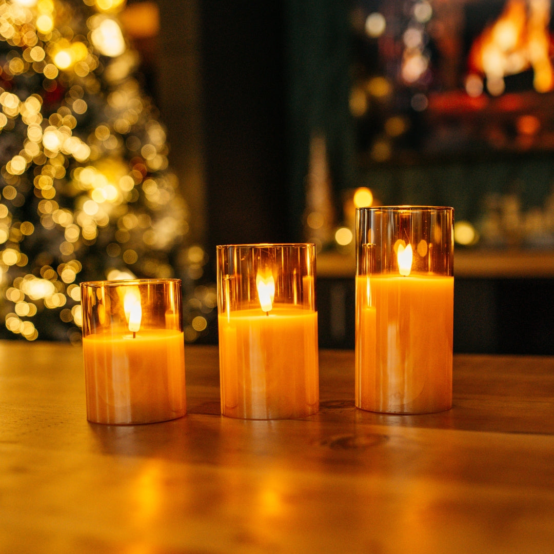 Three lit flameless candles in glass holders on a wooden surface with a blurred Christmas tree and fireplace in the background.