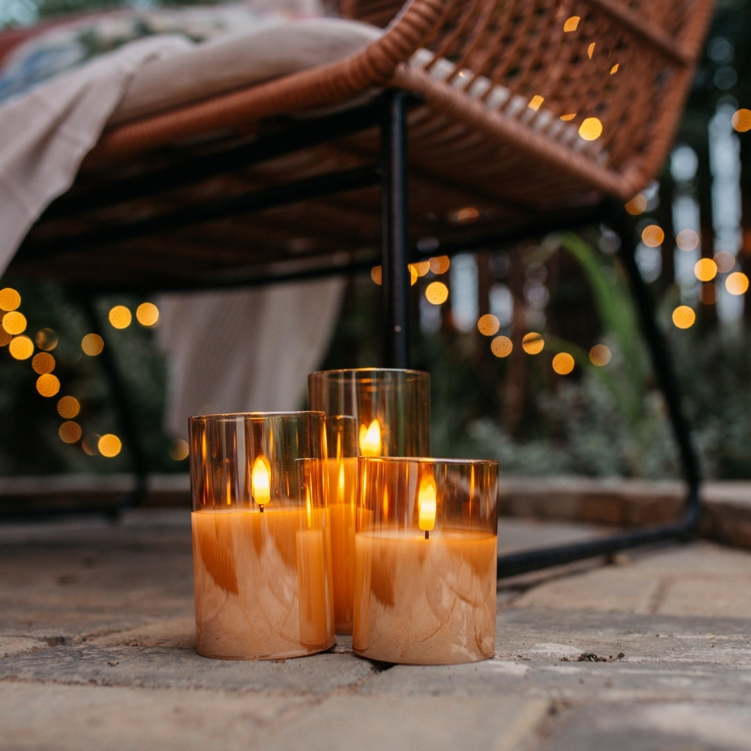 Three lit candles in glass holders on a stone surface with blurred lights and chair in the background
