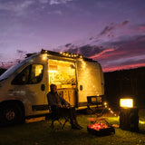 Foldable LED book light glowing beside a campfire outside a camper van under a purple and pink sunset sky