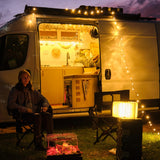 Foldable LED book light glowing beside a woman sitting outside a camper van at night with string lights