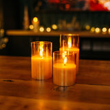 Three lit flameless battery-operated candles in glass holders on a wooden surface with a blurred background
