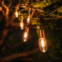 Gold Drop String Lights with warm golden T45 tubular bulbs suspended on individual drop wires on black cable creating a dramatic pendant effect in a styled South African outdoor space at dusk