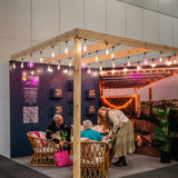 People interacting in a modern indoor setting with wooden beams and gold string lights with pink flamingo bulbs.