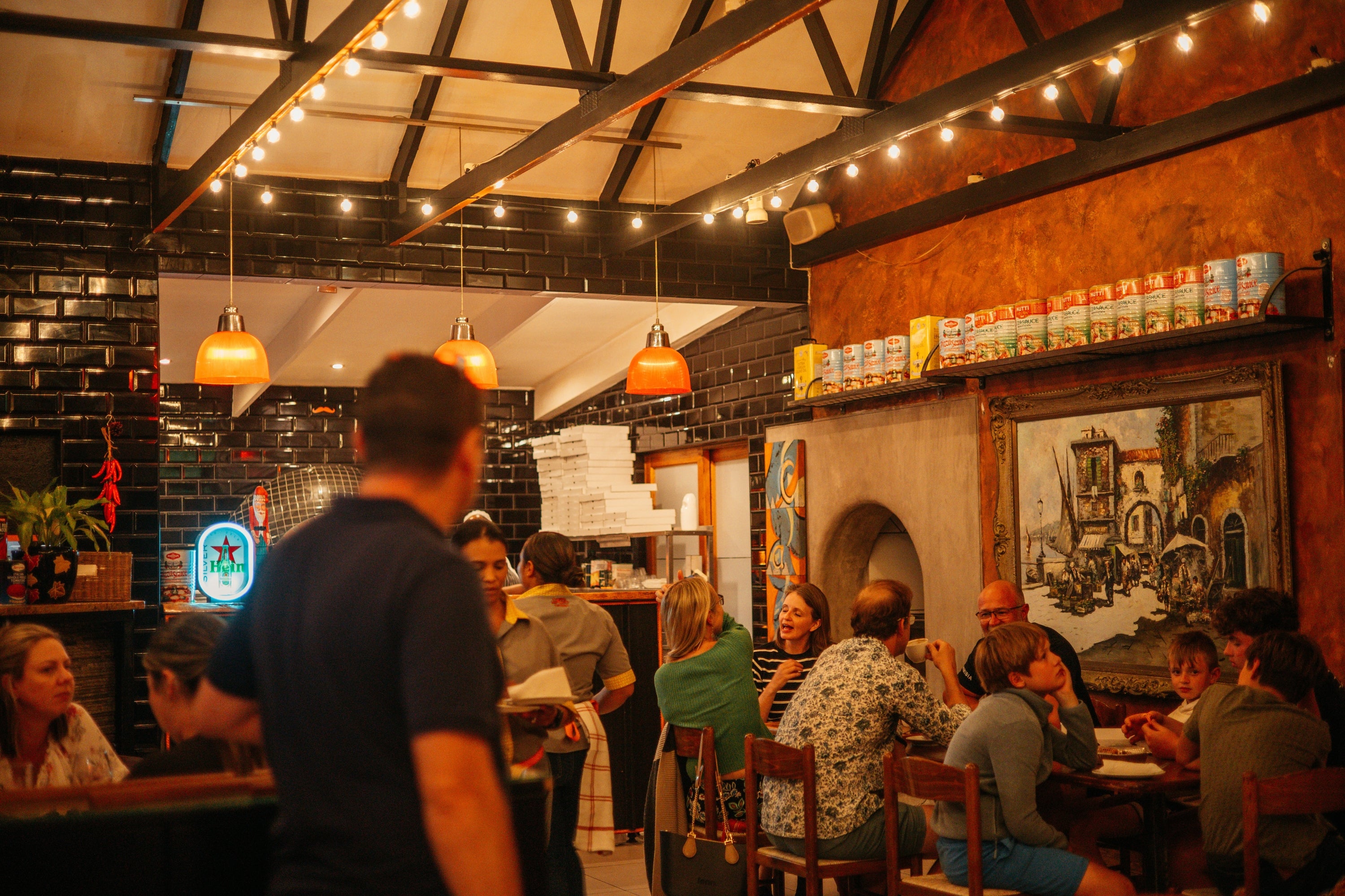 A warm restaurant interior with a group of people dining and socializing around tables under hanging string lights and orange pendant lamps. The space features dark brick and wood walls, a large framed painting, and a tiled counter in the background.