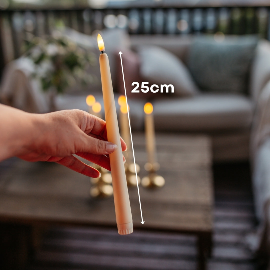 Hand holding ivory flameless taper candle showing 25cm height measurement with candles in gold candlestick holders on outdoor table in background