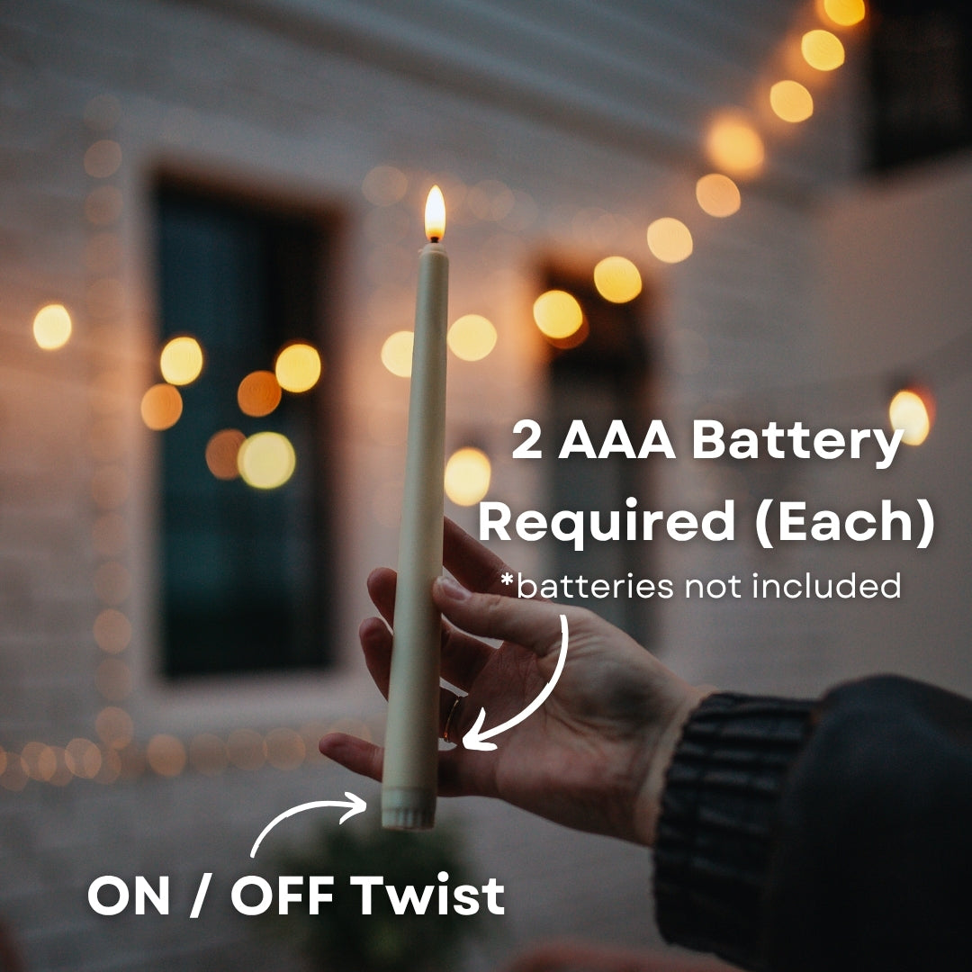 Hand holding ivory flameless taper candle with 2 AAA battery required callout and on/off twist switch indicator with string lights bokeh background