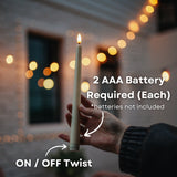 Hand holding ivory flameless taper candle with 2 AAA battery required callout and on/off twist switch indicator with string lights bokeh background
