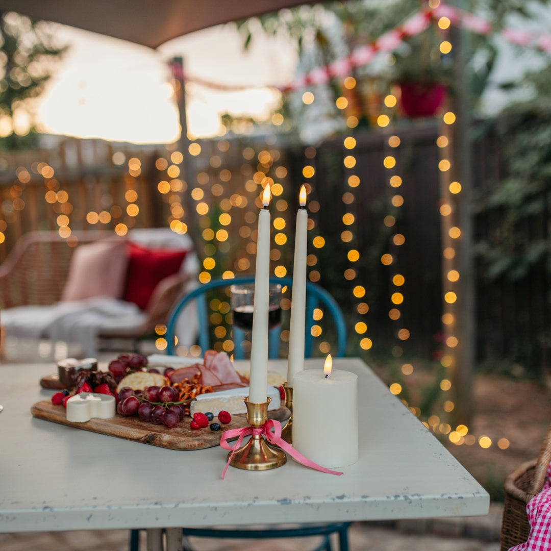 Ivory flameless taper candles glowing on a dinner table with food and fairy lights bokeh in the background