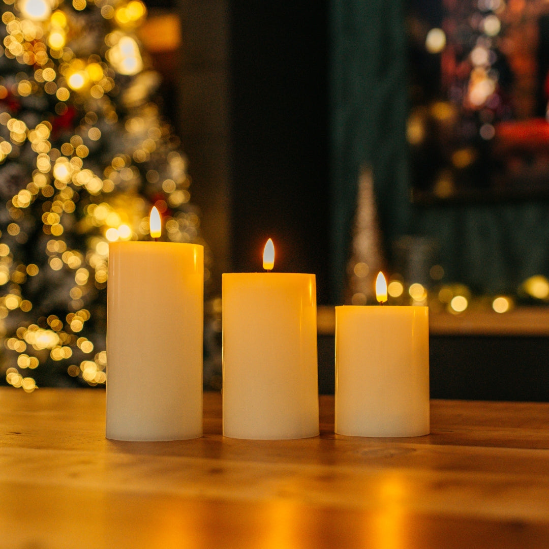 Set of three ivory real wax flameless LED pillar candles glowing on a wooden table with Christmas tree and fireplace bokeh background