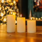 Three ivory real wax flameless LED pillar candles glowing on a wooden table with Christmas tree and fireplace bokeh background