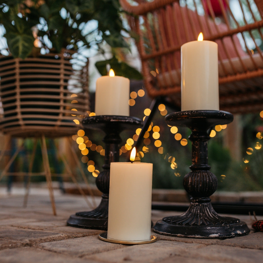 Three ivory real wax flameless LED pillar candles on vintage black candlestick holders outdoors at dusk with fairy lights bokeh background