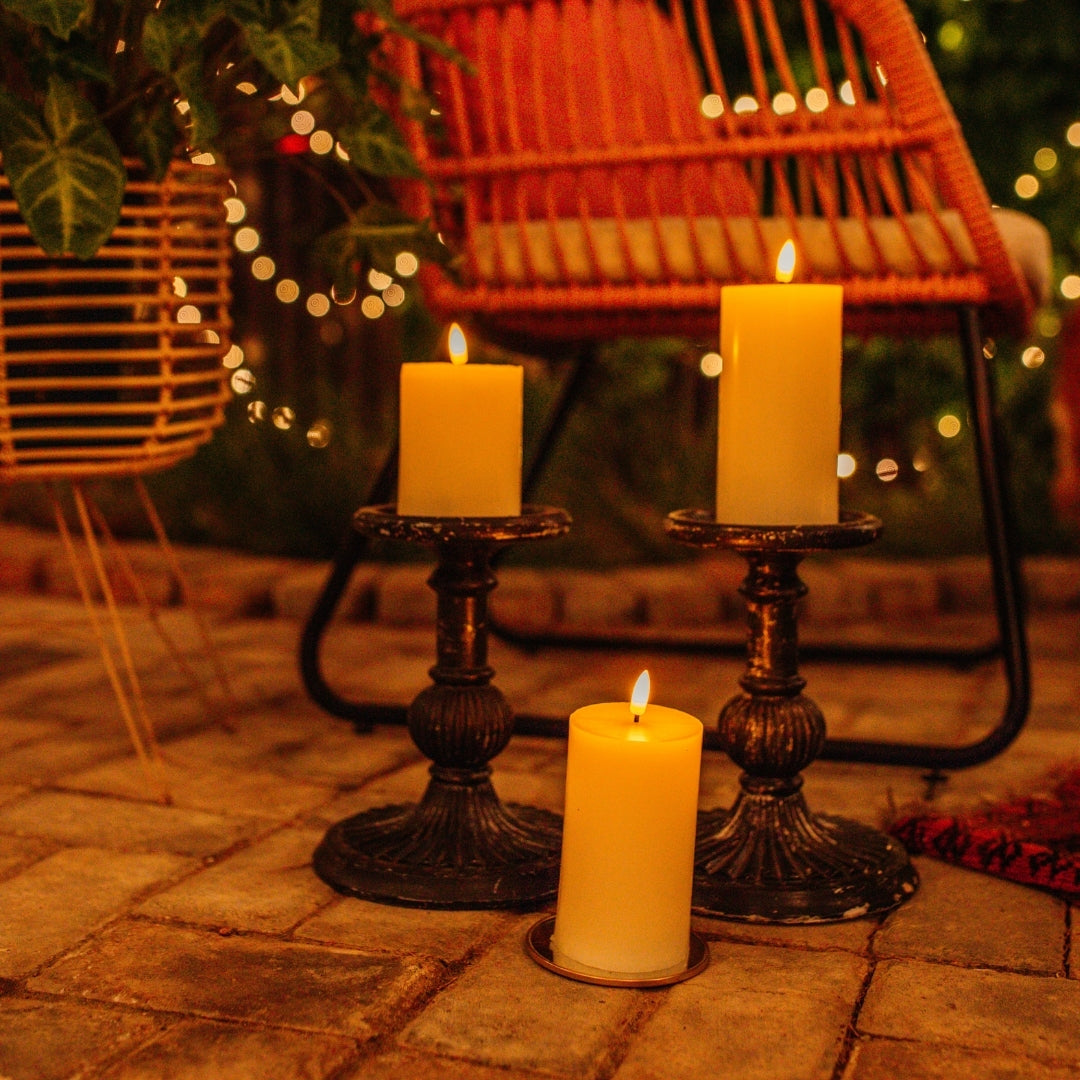 Three ivory real wax flameless LED pillar candles on vintage black candlestick holders on an outdoor patio at night with fairy lights in background