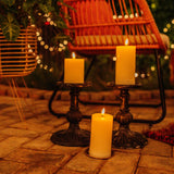 Three ivory real wax flameless LED pillar candles on vintage black candlestick holders on an outdoor patio at night with fairy lights in background