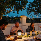Group of friends dining outdoors at dusk with ivory real wax flameless LED pillar candles and greenery as table centrepiece with string lights overhead