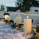 Ivory real wax flameless LED pillar candles arranged as an outdoor dining table centrepiece with greenery garland and string lights at dusk