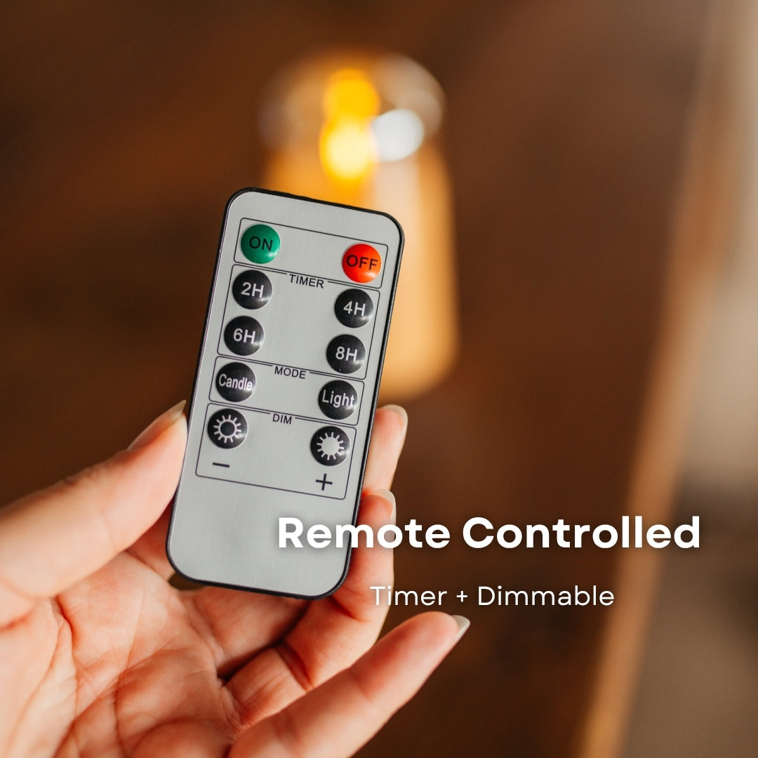 Hand holding remote control for ivory pillar flameless LED candles showing timer and dimming functions with glowing candle in background