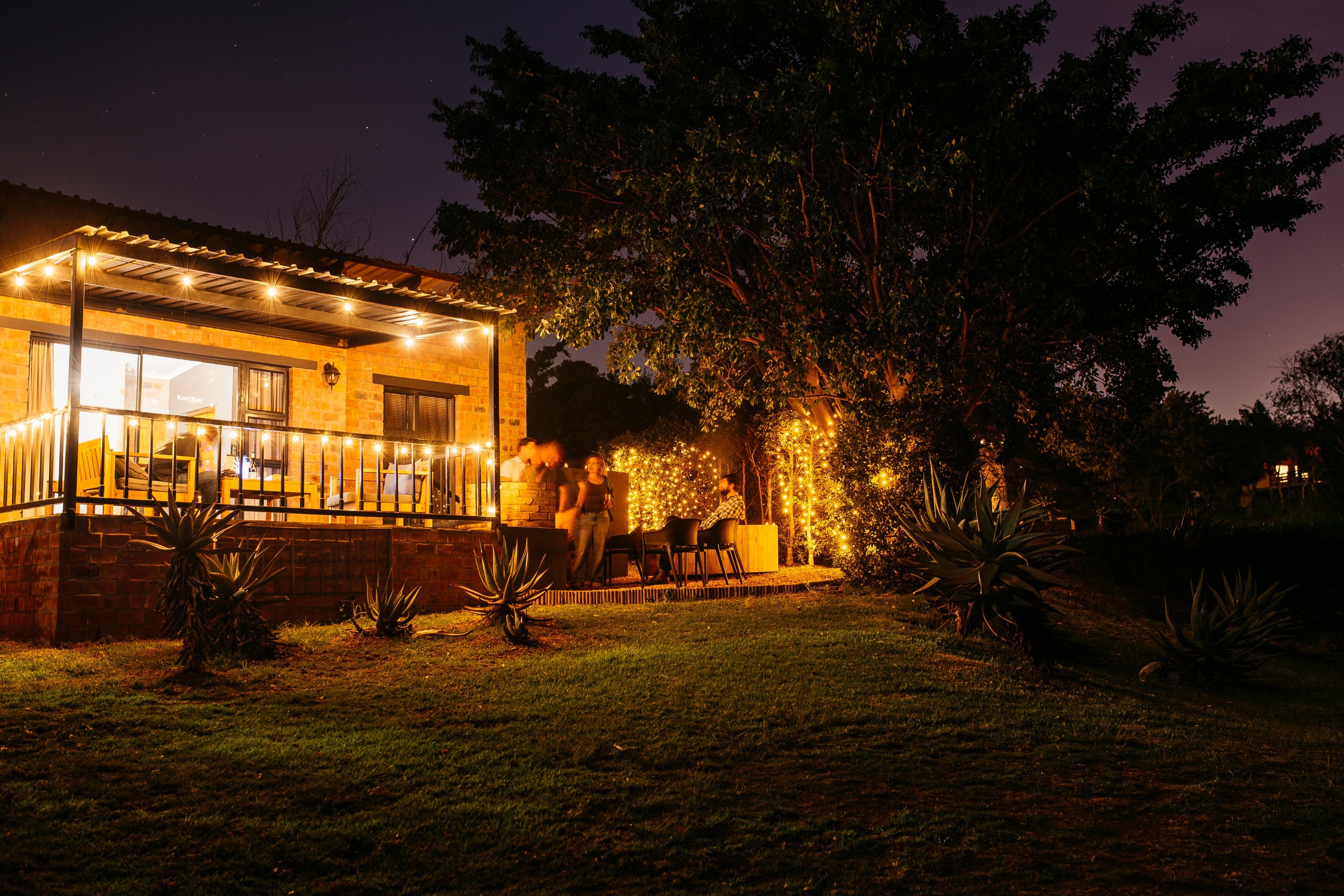 A warmly lit accommodation unit at night with a covered porch, outdoor seating, solar string lights, and a grassy yard with trees in the background.