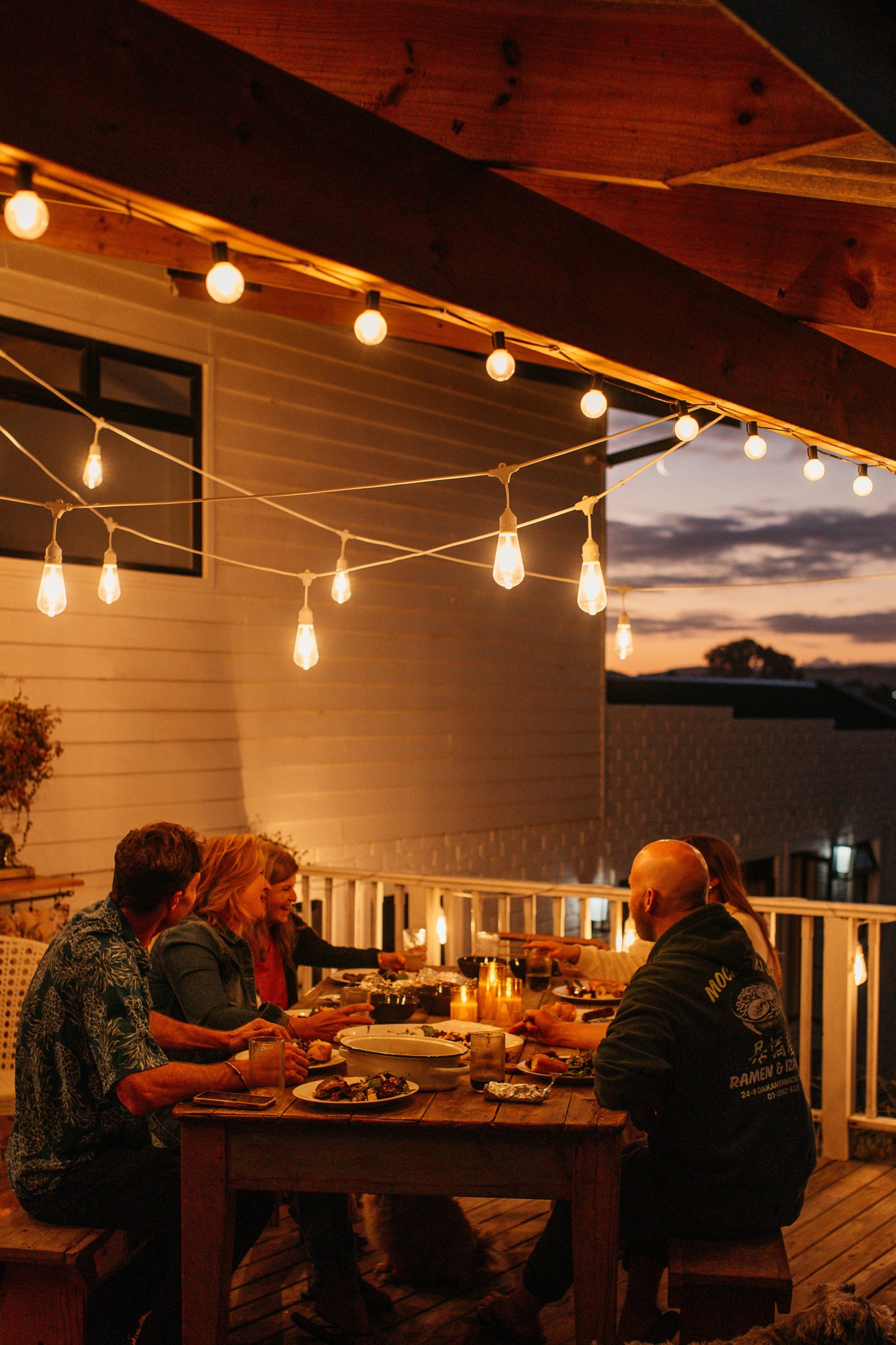People sitting at a table on a patio with warm string lights and a sunset sky.