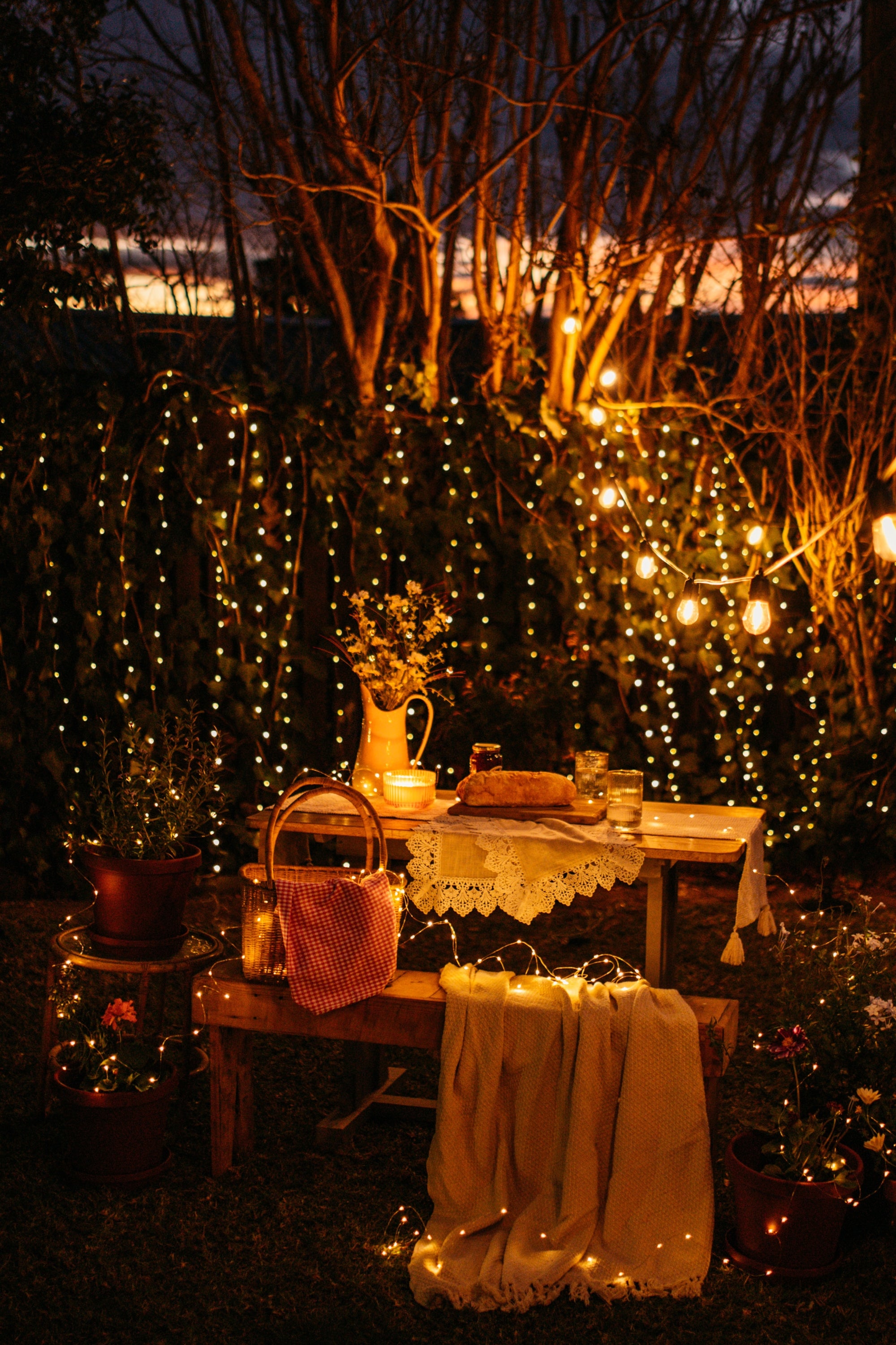 An outdoor evening garden table setup with warm solar string lights and fairy light, a lace tablecloth, candles, a basket, and draped fabric, surrounded by trees and foliage.