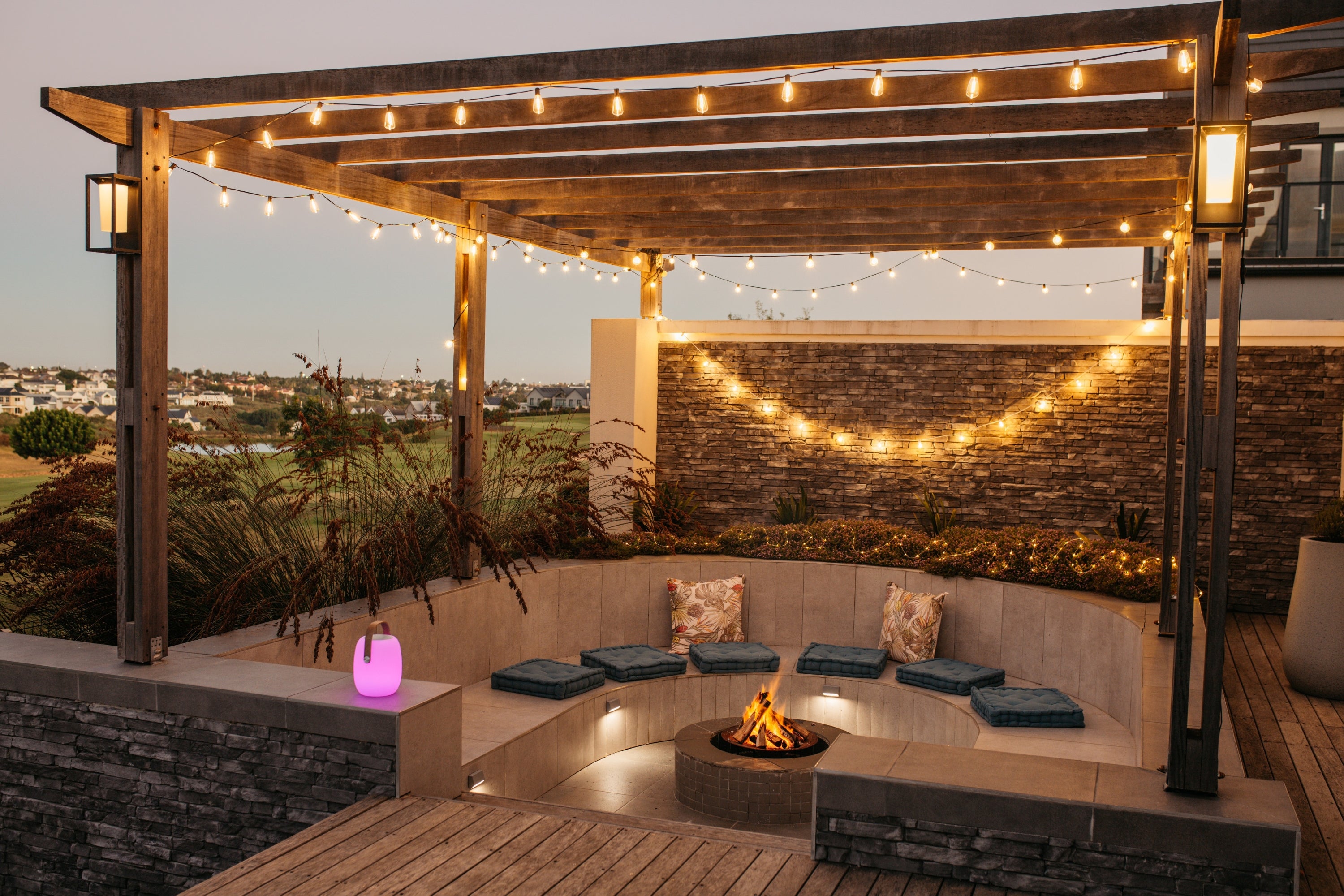 Outdoor patio with wooden pergola, fire pit, warm string lights, and wall lights.