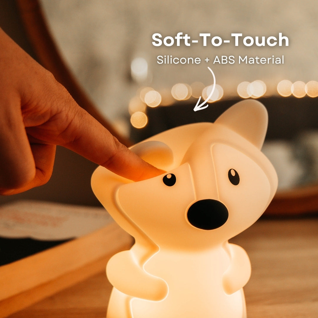 Finger pressing into the soft silicone surface of the Lightie Fox night light glowing warm white with soft to touch and silicone plus ABS material callout