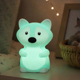 Lightie Fox silicone kids night light glowing teal on a bedside table in a softly lit children's room with fairy lights in the background