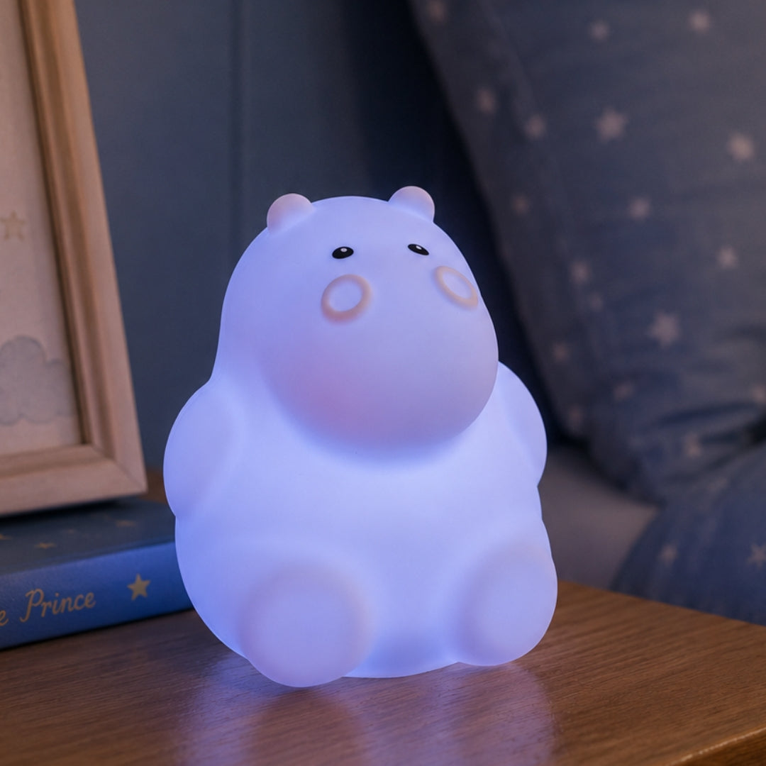 Lightie Hippo silicone kids night light glowing teal on a bedside table in a softly lit children's room with fairy lights in the background