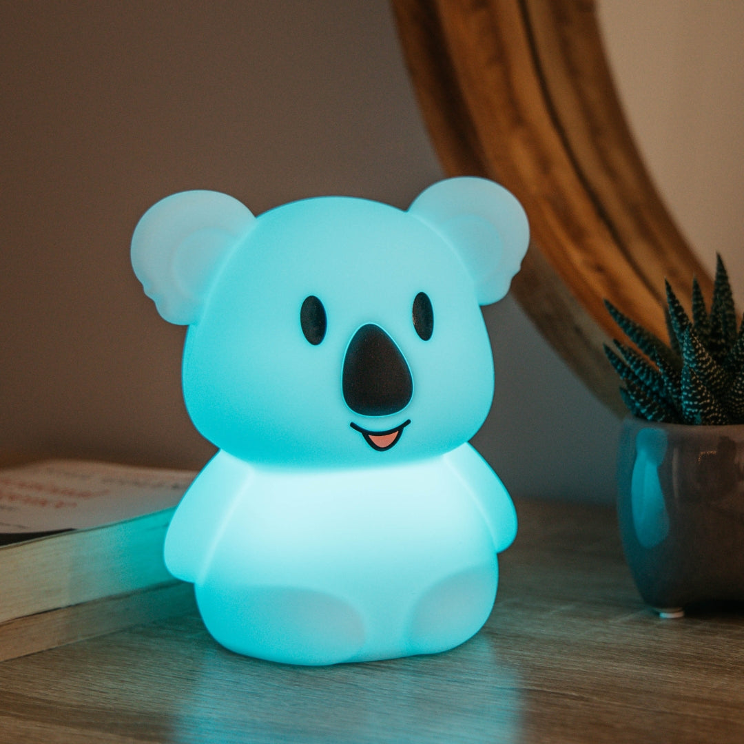 Lightie Koala silicone kids night light glowing teal on a bedside table in a softly lit children's room with fairy lights in the background