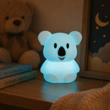 Lightie Koala silicone kids night light glowing warm white on a wooden surface with warm golden bokeh fairy lights in the background