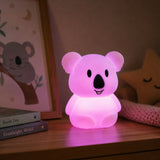 Lightie Koala silicone kids night light glowing warm white on a wooden bedside surface with cushions in the background