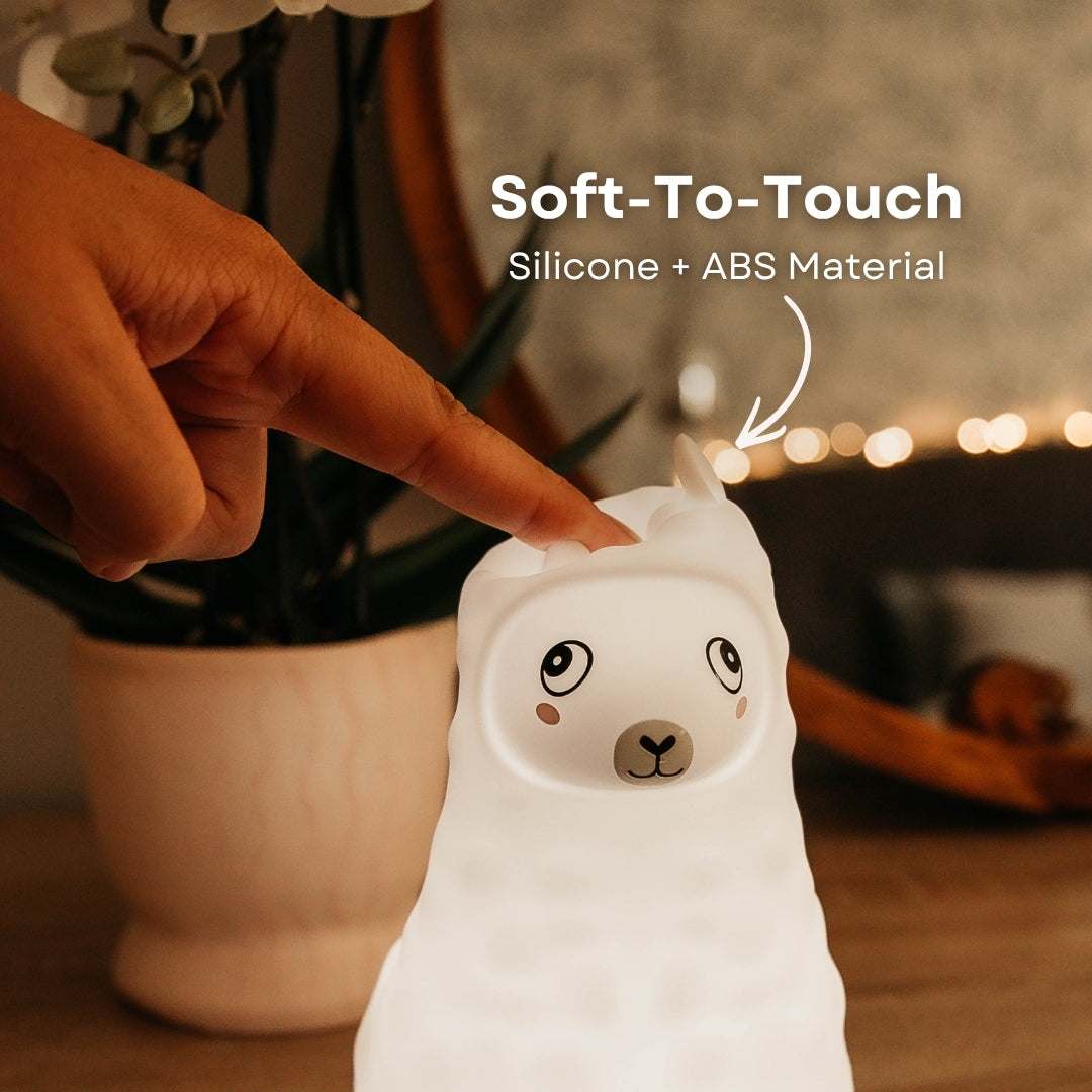 Finger pressing into the soft silicone surface of the Lightie Llama night light glowing warm white with soft to touch and silicone plus ABS material callout